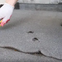 Ręka w rękawicy z nożem przygotowuje dach z papy do naprawy. Widać pęknięcia i dziury, które wymagają interwencji.