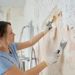 Kobieta w rękawiczkach zeskrobuje starą tapetę szpachelką, przygotowując ścianę do malowania.