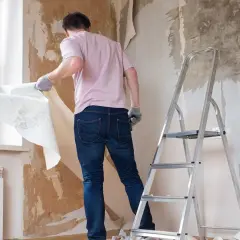 Mężczyzna w rękawiczkach zrywa starą tapetę ze ściany. Obok stoi drabina, przygotowania do remontu.