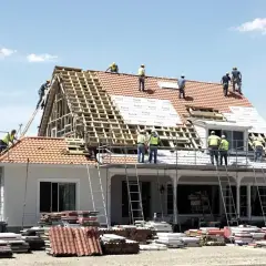Ekipa budowlana pracuje nad wymianą dachu. Widać rusztowania, drabiny i stosy dachówek. Określenie wymiana dachu cena jest kluczowe przy planowaniu remontu.