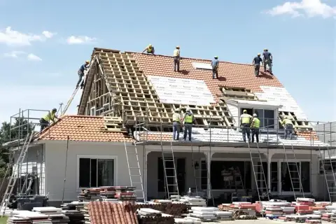 Ekipa budowlana pracuje nad wymianą dachu. Widać rusztowania, drabiny i stosy dachówek. Określenie wymiana dachu cena jest kluczowe przy planowaniu remontu.