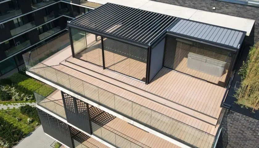 Nowoczesny taras na dachu z przeszklonym pawilonem i pergolą. Drewniana podłoga i szklane balustrady tworzą elegancką przestrzeń.