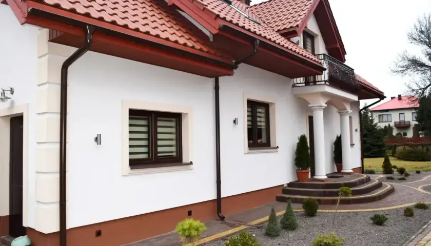 Nowoczesny projekt elewacji starego domu z białym tynkiem, brązowymi okiennicami i czerwoną dachówką.