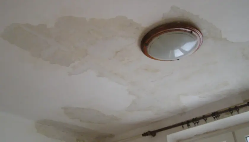 Plamy na suficie świadczą o jego higroskopijności i problemach z wilgocią. Widoczna lampa sufitowa i fragment okna.